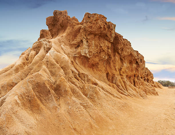 A dramatic sandstone structure exhibits weathered textures and detailed erosion patterns. The rich orange and brown tones highlight the natural beauty of this rock's surface, set against a pastel-toned sky.