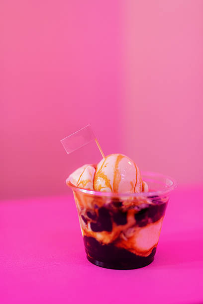 A close-up shot of a whimsical dessert in a clear plastic cup. The dessert features layers of boba pearls at the bottom, creamy vanilla ice cream, and a drizzle of caramel sauce on top. A small pink flag on a toothpick adds a playful touch, all set against a soft, pink-hued blurred background suggesting a fun, festive atmosphere.