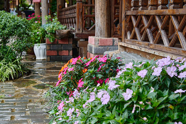 Wet stone pathway winding past vibrant impatiens and a traditional wooden porch railing