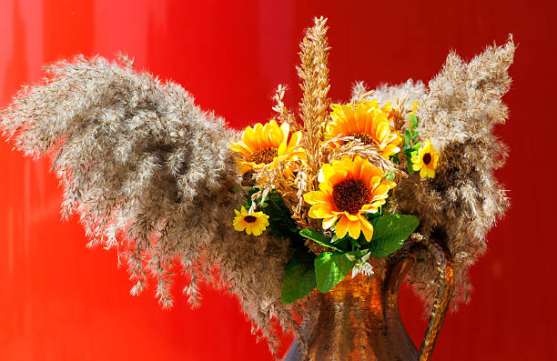 A vase with flowers and a plant in it. The vase is made of metal and is placed on a red background