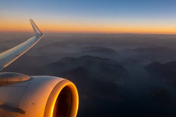 Inspirational view from an airplane window during sunrise, featuring the aircraft wing and jet engine glowing in golden morning light. Below, misty mountains and a winding lake create a breathtaking aerial landscape. Perfect for themes of air travel, wanderlust, adventure, freedom, modern transportation, and global tourism.