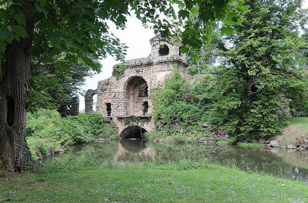 Old stone ruins stand by a quiet pond, their reflection visible in the calm water surrounded by lush green trees.