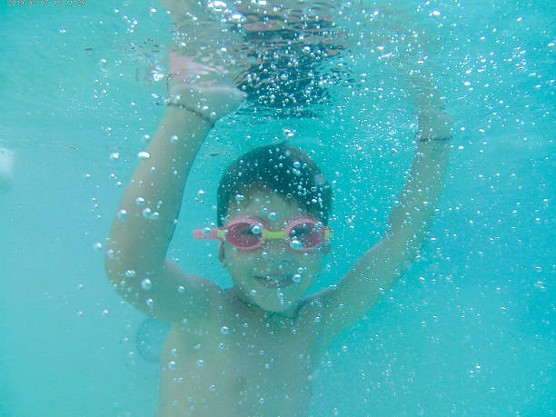 Child wearing goggles swimming underwater in a bright blue pool, looking at the camera and surrounded by bubbles