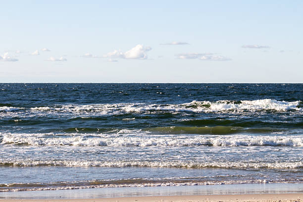 Waves roll onto a sandy beach, creating foamy edges as they crash on the shore. The clear blue sky is dotted with a few clouds, suggesting a pleasant early afternoon.