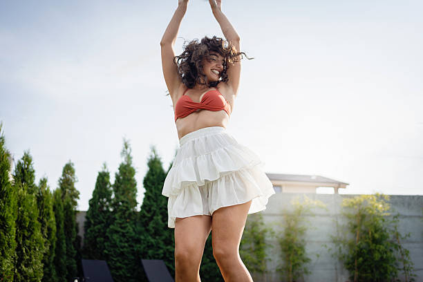 A carefree woman in cheerful motion next to a well-maintained pool in an outdoor setting, exuding happiness and summertime relaxation.