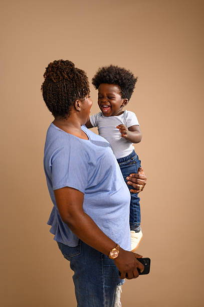 Smiling mother holds her young son who laughs joyfully with eyes closed. Both dressed casually in jeans and shirt against plain brown studio background.