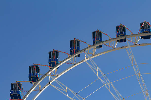 Bright blue cabins of a ferris wheel rise high in the sky, showcasing a fun atmosphere at an amusement park on a sunny day. Excitement fills the air as people enjoy the view.