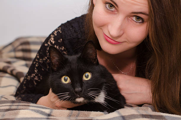 Young woman with long brown hair lies on plaid blanket beside black cat with white patch. Both share calm moment, eyes directed at camera, soft indoor light enhancing scene