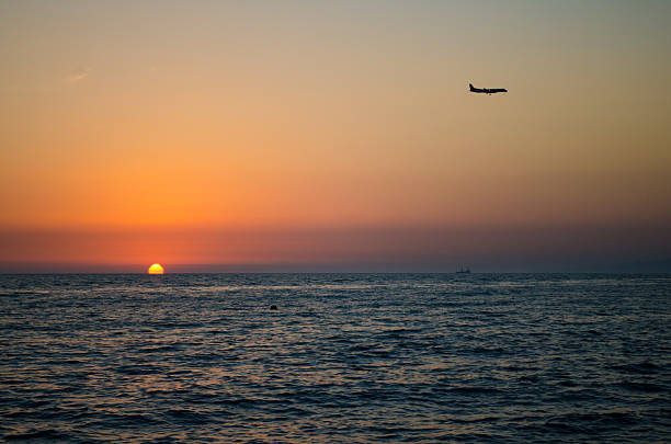 Airplane glides above calm ocean, silhouetted against vibrant sunset. Orange and purple tones dominate sky, merging with darkening sea. Peaceful, serene atmosphere captured from shoreline