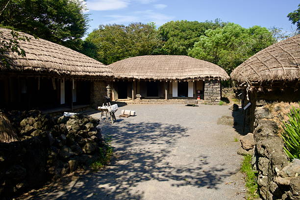 A broad courtyard is enclosed by connected thatched houses at Jeju Folk Village, with work benches and tools placed under deep eaves.