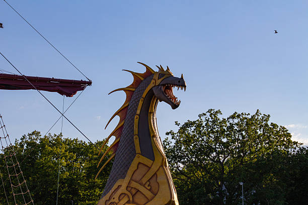 A large dragon figure stands tall in a public park during a sunny evening. The figure has vibrant colors and intricate designs, while the sky is clear with a few scattered clouds.