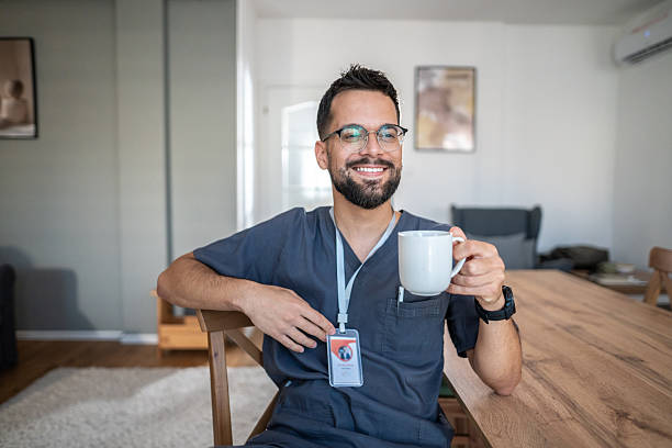 Male healthcare professional in scrubs and id badge sits at wooden table, smiling and relaxed while holding a mug of coffee during a peaceful work break at home-like setting