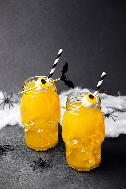 Two spooky skull glasses hold yellow drinks decorated with candy eyes and bat picks. Cobwebs and spiders add Halloween atmosphere.