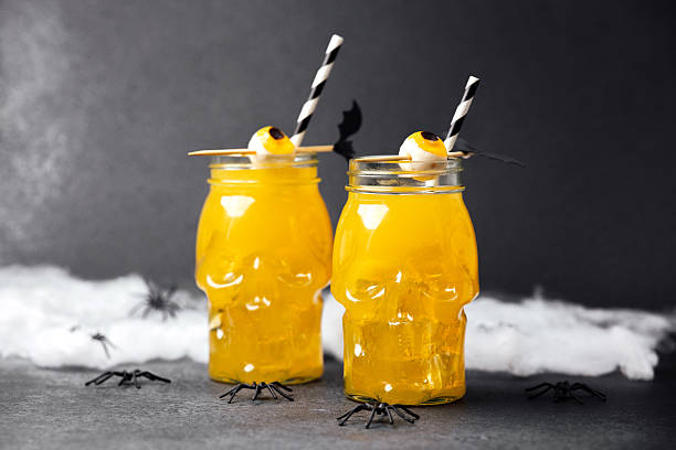 istock Two skull glasses filled with yellow drink, ice, straws, and candy eyeballs on a dark surface with cobwebs and spiders 2236710805