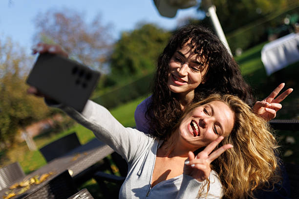 Two joyful friends capture a fun moment together in a park, surrounded by lush greenery and outdoor seating. They pose with peace signs and big smiles in the sunlight.