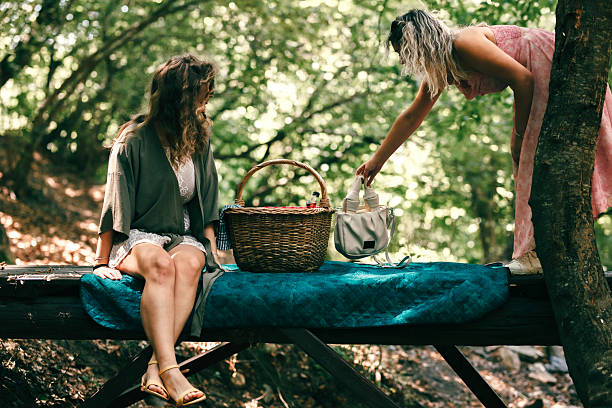 Two friends gather at a peaceful spot in the forest, sitting on a wooden platform with a picnic basket and sharing drinks. The sunlight filters through the trees in this tranquil setting.