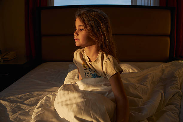 Young girl sitting in bed, looking thoughtfully into golden morning light