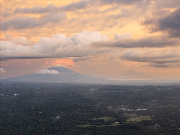 Sunset casts warm glow over distant mountains and green valley, with clouds creating dynamic sky.
