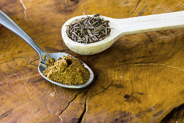 Two different types of teaspoons, one filled with cumin seeds and one filled with ground cumin powder on a wooden background.