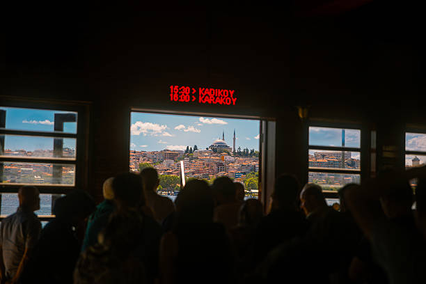A group of passengers waits inside a ferry terminal in Istanbul, framed by the glowing digital timetable showing departures to Kadıköy and Karaköy. Through the large windows, the city’s skyline unfolds with a prominent mosque and the colorful hillside houses. The image captures the rhythm of urban transit, blending tradition with modernity. Ideal for illustrating Istanbul’s daily life, transport system, and the connection between the city’s European and Asian sides.