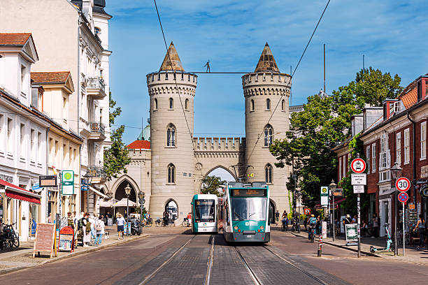 Vibrant urban scene in Potsdam, Germany, featuring modern trams on a busy street framed by the medieval gate towers. A lively mix of tourists and locals under a clear blue sky.