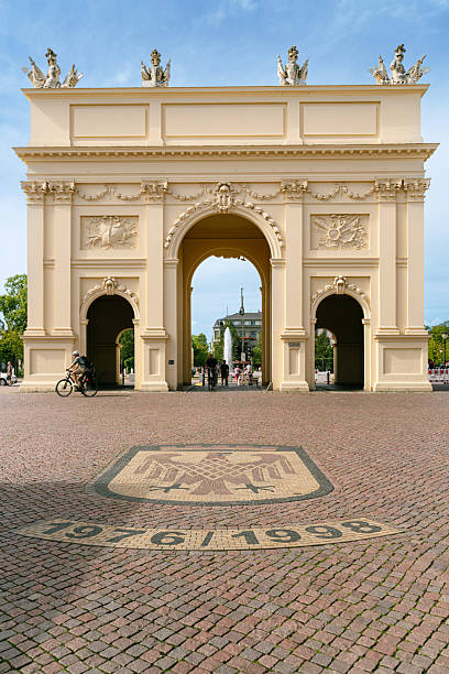 A grand neoclassical triumphal arch frames a bustling square in Potsdam, Germany. Beam of blue sky, pedestrian activity, and decorative bas-reliefs create a timeless city landmark atmosphere.