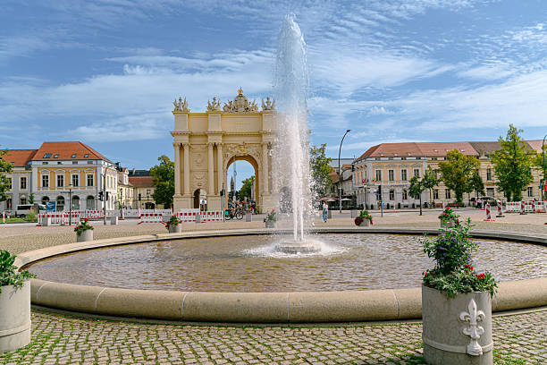 Sunlit Potsdam square features a grand cream arch gateway, a tall central fountain, and surrounding historic buildings. People stroll under a clear blue sky, capturing European charm and civic atmosphere.