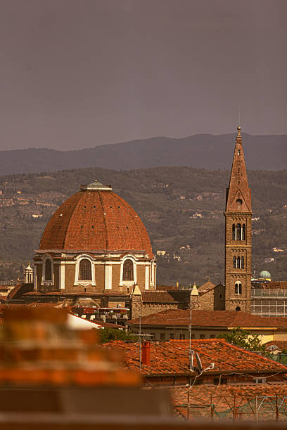 In the photo, we see an urban landscape of Florence (Italy). Prominently featured in the foreground is a large red-tiled dome in Renaissance style, accompanied by a tall, slender stone bell tower. Both stand out among the reddish rooftops of the city, with the Tuscan hills fading into the background. The scene reflects the classic and charming character of Florence, known as the birthplace of the Renaissance