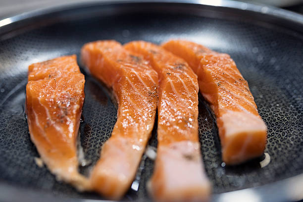 Four fresh salmon fillets arranged side by side in a non-stick frying pan, lightly seasoned and glistening with oil. The pan’s textured surface hints at even heat distribution, while the fillets—rich in color and marbled with fat—suggest quality and care in preparation. Shot with Canon R5
