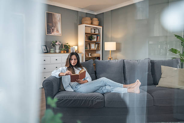 Young adult woman relaxing on a comfortable gray sofa in a stylish living room while reading a book, enjoying a peaceful leisure moment in a calm indoor atmosphere