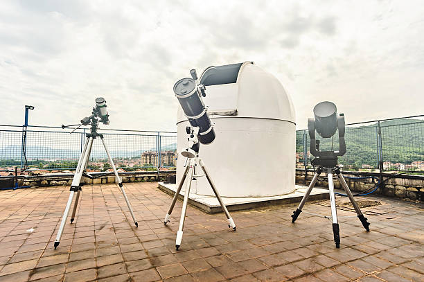 A rooftop observatory setup featuring multiple telescopes on tripods around a white dome, against a cloudy sky and distant landscape. Emphasizes science, exploration, and astronomical observation