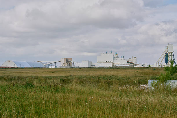 Main processing building of a potash plant in Saskatchewan, Canada, with white industrial architecture, ventilation stacks, and smoke rising under dramatic dark clouds.