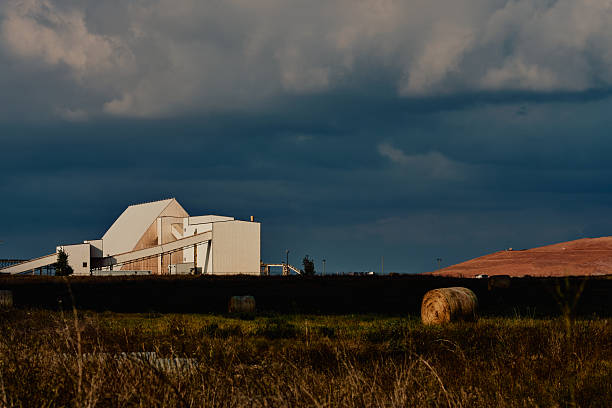 Industrial potash facility in Saskatchewan, Canada, with white processing buildings, conveyor systems, hay bales in the foreground, and tailings pile under dramatic cloudy sky.