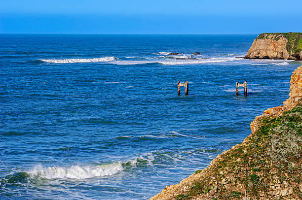 Ocean waves crashing along rocky shore of Davenport, with remnants of the old concrete Davenport Pier, viewable in the far distance.Taken in Davenport, California, USA.