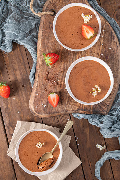 istock Three white bowls of rich chocolate mousse, one with a golden spoon, are elegantly arranged on a wooden board and table with fresh strawberries and delicate white flowers 2236813954
