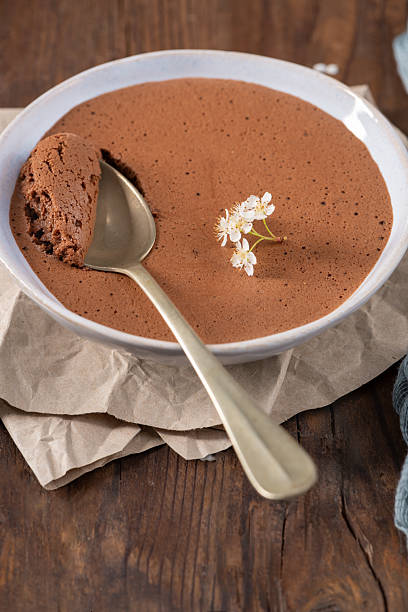 istock Velvety chocolate mousse in a white bowl, a golden spoon scooping a portion, adorned with delicate white flowers, set on crumpled parchment paper over a rustic wooden table 2236813983