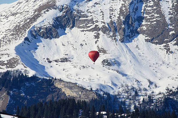 Red hot air balloon floating above snow-covered alpine peaks with pine forest below. Winter adventure, travel, and scenic mountain landscape.