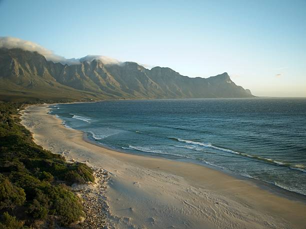 The attached image captures a serene coastal landscape with a stunning combination of ocean and mountains. A wide stretch of sandy beach runs along the shoreline, bordered by lush green vegetation in the foreground. The ocean appears calm, with gentle waves meeting the sand, while the water reflects different shades of blue. In the background, a majestic mountain range rises steeply, partially draped with soft clouds near the peaks. The sky is clear and bright, suggesting it’s either early morning or late afternoon, adding a peaceful and refreshing feel to the scene.