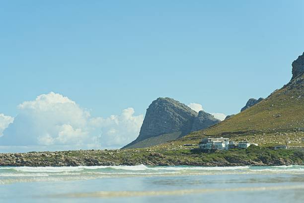 A scenic photograph shows a cluster of modern, white buildings nestled into the base of a large, rocky mountain. The buildings stand on a grassy, elevated point along the coastline. The foreground is a blurred shot of a sandy beach, with gentle waves rolling in. A low-lying rocky breakwater separates the land from the sea. The sky above is a clear, serene blue with a few fluffy white clouds. The entire scene is framed by the steep slope of the mountain on the right and the vast expanse of the ocean.