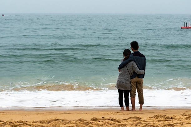 Two Young Asian men standing  on the beach, hug each other neck, point at the edge of sea with fun, cheerful, smile of happiness and hopefulness. Two men being with hope at beach concept at sunset time.