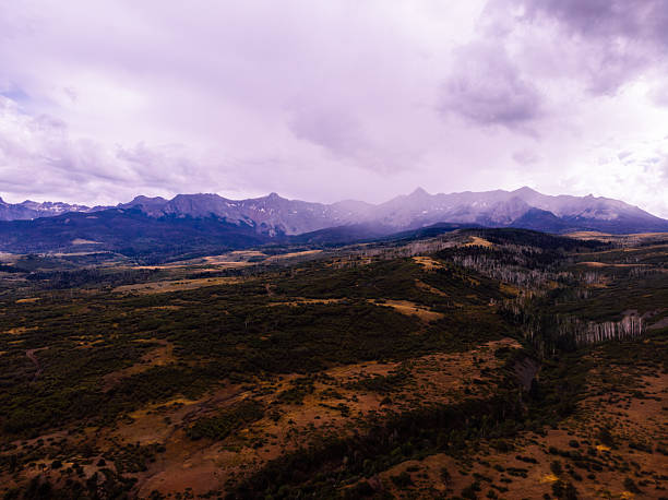 Expansive aerial view capturing the diverse landscape of Ouray County, Colorado. Terrain transitions from rocky foreground to distant mountain ranges with purple-tinted cloudy sky. Landscape features mix of brown earth tones, dark vegetation patches, and dramatic mountain silhouettes in background.