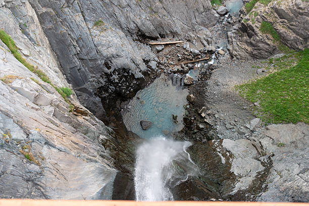 A narrow waterfall flows between steep rocky walls with moss-covered stones. Water creates a misty cascade through a tight rocky channel with green vegetation visible on the edges.
