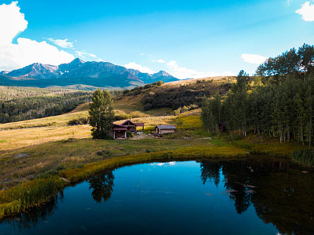 Two rustic wooden cabins situated in a scenic valley with a reflective pond in the foreground. Snow-capped mountain peaks rise in the background, surrounded by aspen and pine forests. Golden grasslands and green vegetation create a vibrant landscape in San Miguel County, Colorado, showcasing the region's natural beauty during late summer or early autumn.