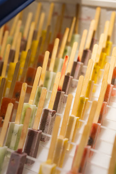 close-up shot of a large display of numerous popsicles, each on a wooden stick. The popsicles are a variety of vibrant colors and flavors—including red, green, yellow, and chocolate—arranged in a neat grid on a white display rack.