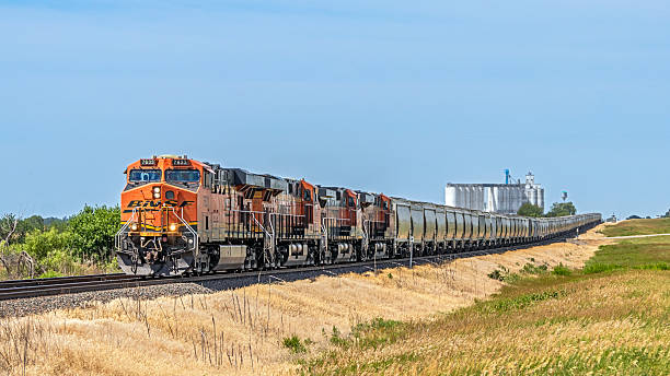 Walton KS, United States - June 16, 2018: In rural Kansas, a long and heavy BNSF grain train with four diesel locomotives hard at work, passes a huge grain storage facility (silos/elevators) in the background. Wide angle view.