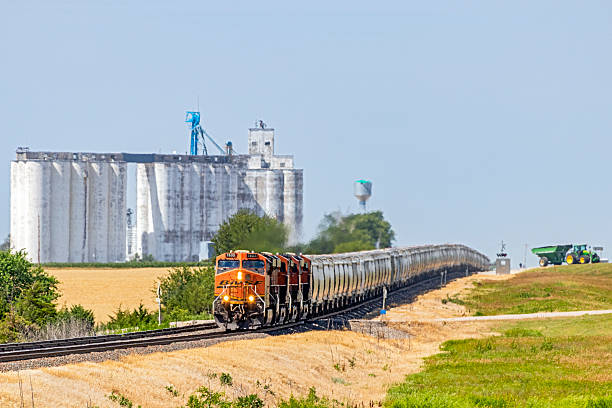 Walton KS, United States - June 16, 2018: In rural Kansas, three agricultural themes in one frame: a long BNSF Grain Train; in the background a huge grain storage facility (Silos/elevators); and a grain farmer departing the facility in his tractor and trailer.