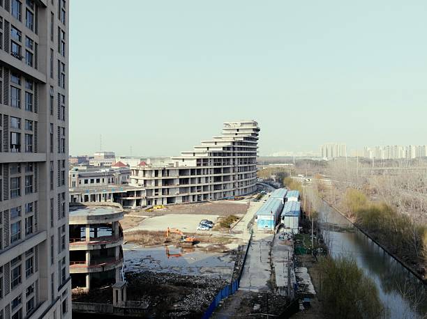 Captivate your audience with this compelling view of a bustling urban construction site, featuring a distinctive tiered or stepped high-rise building under construction, surrounded by excavators, temporary structures, debris, and a serene river channel, with distant city skylines and hazy horizons under a clear sky. This dynamic scene highlights modern architectural innovation and urban growth, ideal for real estate promotions, engineering visuals, city planning designs, or content showcasing infrastructure development, construction progress, and metropolitan expansion!
