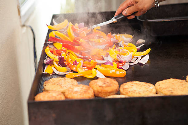 A close-up shot captures an unrecognizable cook, wearing a blue glove, using a spatula to flip a hamburger patty on a large, hot griddle.