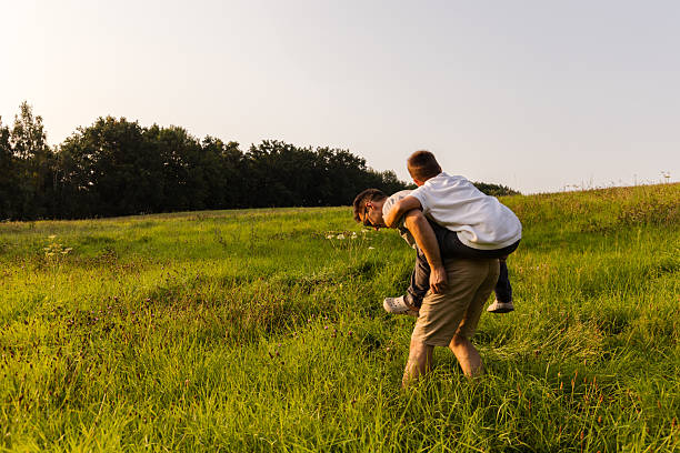 A Caucasian father carries his smiling daughter on his shoulders in a scenic meadow at sunset, surrounded by lush greenery and distant mountains, creating a warm and serene outdoor setting.