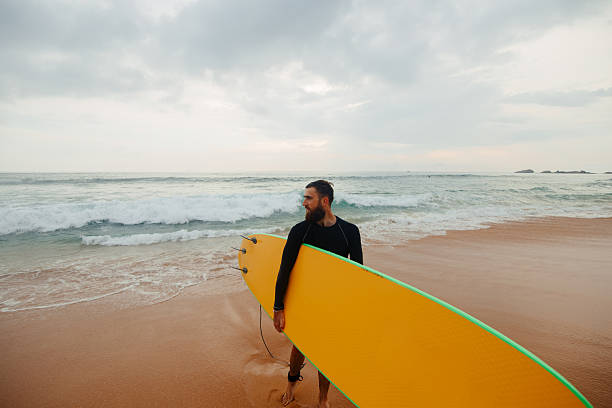 istock Young  bearded man with surfboard standing near a beach. Man with surfing board outdoors on a summer day. 2236893515